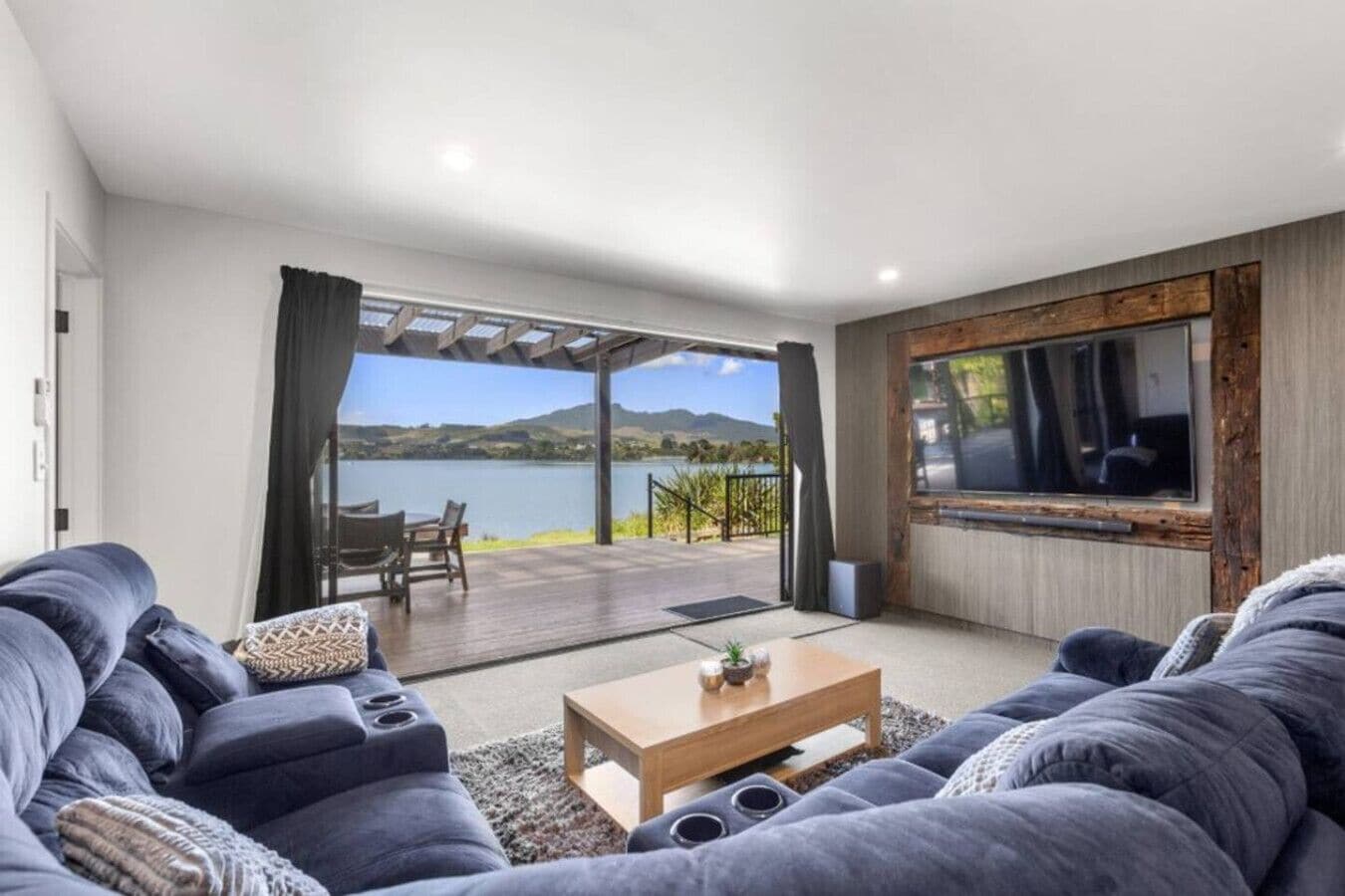 A modern living room with a large sofa, a coffee table, and a TV. There is a patio area with a view of a lake and hills.