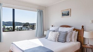 A bedroom with a large window, a bed with multiple pillows, a bedside table with a lamp, and a framed picture on the wall. - Harbourside Cove • Views, Cosy + Peaceful (Raglan)
