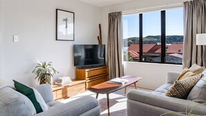 A modern living room with a flat-screen TV, two sofas, a coffee table, and a large window with a view of buildings. - Harbourside Cove • Views, Cosy + Peaceful (Raglan)