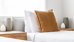 Ein ordentlich bezogenes Bett mit weißer Bettwäsche, einem braunen Kissen und einem grauen Kissen. Auf den Holz-Nachttischen neben dem Bett stehen zwei weiße Vasen.
