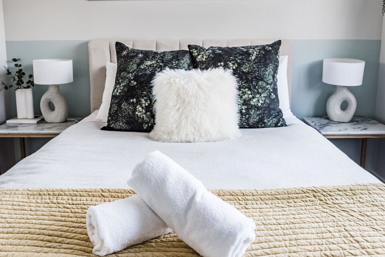A neatly made bed with a white duvet, two black pillows with a floral pattern, a fluffy white pillow, and a beige textured throw blanket.