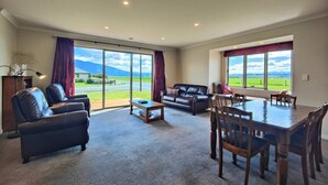 A spacious living room with a large window, a sofa, a coffee table, and a dining table with chairs. - Fiordland Family Favourite (Te Anau)