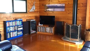 Interior acogedor de una cabaña con paredes de madera, un televisor, una estantería, una chimenea y una ventana con vistas a las montañas.