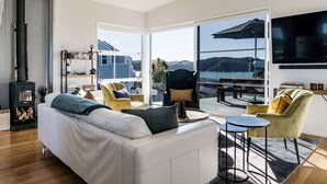A modern living room with a white sofa, two armchairs, a coffee table, and a fireplace. Large windows offer a view of a lake and hills. - Government House • Spa • Comfort • Views (Raglan)