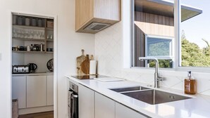 A modern kitchen with white cabinetry, a double sink, and a window providing a view of greenery outside. - Government House • Spa • Comfort • Views (Raglan)