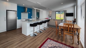 A modern kitchen with a central island, bar stools, a dining table with chairs, and a large window with a view of greenery. - Rack n Relax Riverton (Riverton / Aparima)