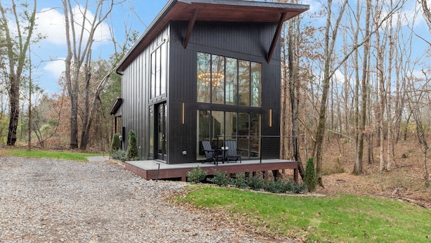 A modern cabin with a large glass window, a wooden deck, and a gravel driveway. - Modern Black Cabin w/ Hot Tub (Burns)