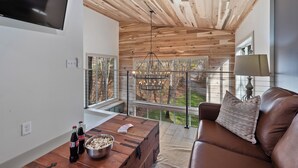 A modern living room with a wooden ceiling, a leather sofa, a coffee table with bottles and popcorn, and a view through large windows. - Modern Black Cabin w/ Hot Tub (Burns)