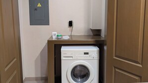 A compact laundry room with a Bosch washing machine, a power outlet, and a small shelf with a cup and papers.