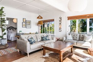 Ein geräumiges Wohnzimmer mit grauem Sofagarnitur, einem Holztisch und einem großen Fenster mit Blick auf Palmen.