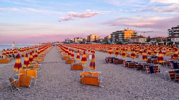Ein Strand mit Reihen von Liegen und Sonnenschirmen, Gebäude im Hintergrund und einem klaren Himmel.