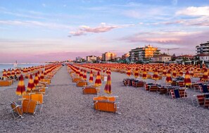 Una spiaggia con file di lettini e ombrelloni, edifici sullo sfondo e un cielo sereno.