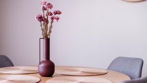 A round wooden table with a dark vase containing pink flowers, surrounded by woven placemats. - Park Podolski Deluxe Apartment (Łódź)