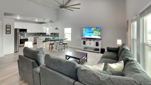 A modern living room with a grey sectional sofa, a coffee table, and a TV on a stand. There is a kitchen in the background with white cabinets and a dining area with a table and chairs. - Coastin' (Rockport)
