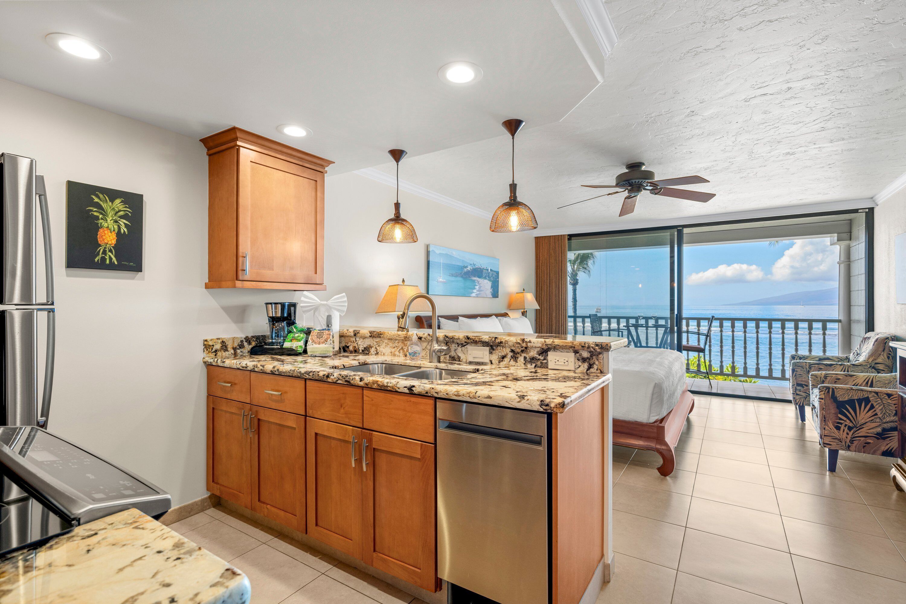 A kitchen with wooden cabinets, a dishwasher, and granite countertops. There is a dining area with a table and chairs, and a balcony with a view of the ocean.
