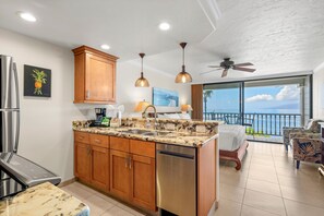 A kitchen with wooden cabinets, a dishwasher, and granite countertops. There is a dining area with a table and chairs, and a balcony with a view of the ocean. - New Listing! Lahaina Shores 309: Oceanfront Studio (Lahaina)