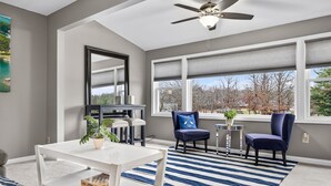 A modern living room with a white coffee table, two blue chairs, a ceiling fan, and a large window with blinds.