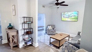 Een woonkamer met een bank, salontafel en plafondventilator. Er is een boekenkast en een bijzettafeltje met een vaas.
