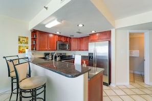 A kitchen with wooden cabinets, a stainless steel refrigerator, a microwave, and a granite countertop. - Corner Unit Beachfront with Secluded Beach! (Destin)
