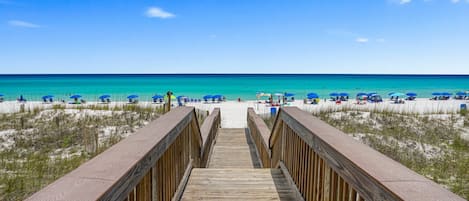 A wooden boardwalk leading to a beach with blue umbrellas and people.