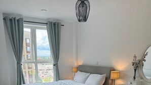 A bedroom with a bed, a desk, a chair, and a large window with curtains. - Barking Bliss - Bright Apartment With City Views (Barking)