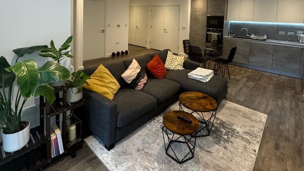 A modern living room with a grey sectional sofa, a coffee table, and a dining area with chairs. There is a kitchen in the background with a sink and cabinets. - Barking Bliss - Bright Apartment With City Views (Barking)