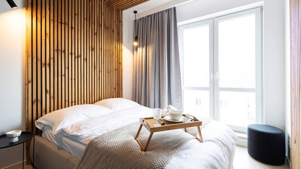 A modern hotel room with a wooden slatted headboard, a large window with curtains, a bed with white linens, and a bedside table with a tray. - Humo Pozna Apartament z Klimatyzacj Gara em (Poznań)