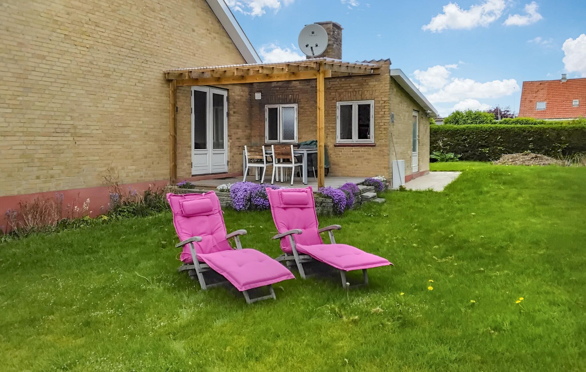 Een huis met een bakstenen gevel, een overdekte patio met een tafel en stoelen, en twee roze loungestoelen in de tuin.