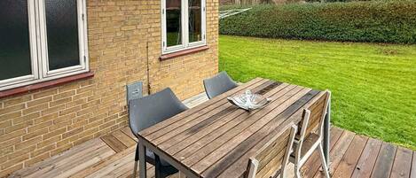Une terrasse en bois avec une table et des chaises, un mur en briques et une pelouse bien entretenue.