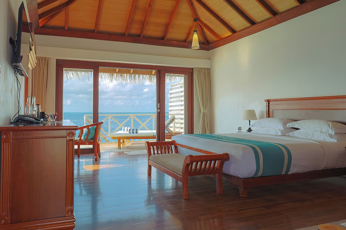 Habitación de hotel con techo de madera, cama con ropa blanca, escritorio con teléfono y vista al mar a través de una puerta de cristal.