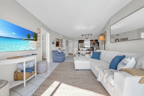 A modern living room with a large TV displaying a beach scene, a white sofa with blue cushions, a dining area with a white table and chairs, and a kitchen area with a hanging light fixture. - Island Oasis: Huge Pool/Spa/Tennis, 1/1 sleeps 6 (Marco Island)