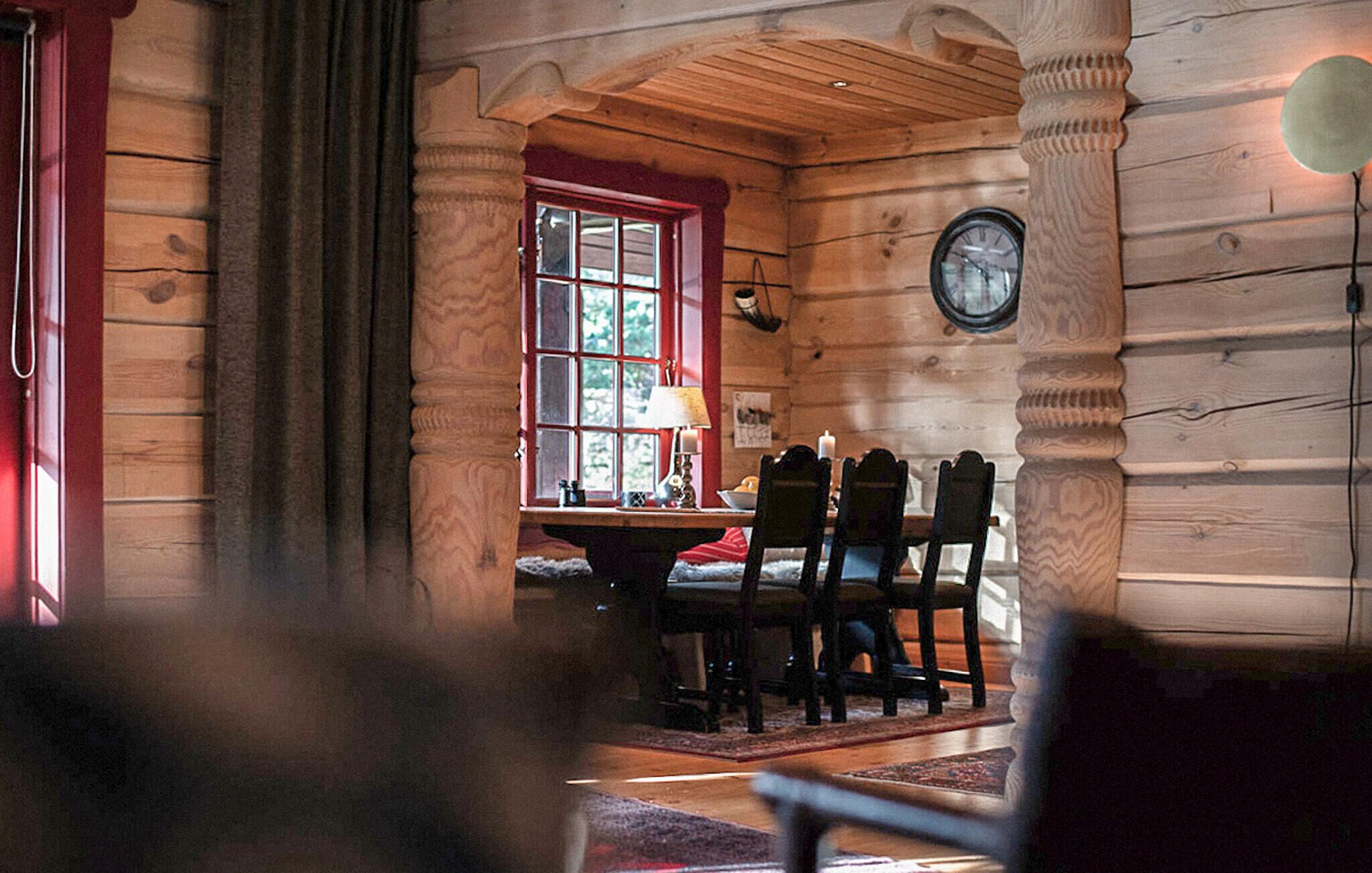Ein Holzhütten-Interieur mit Essbereich, inklusive Tisch, Stühlen, einem Fenster mit rotem Rahmen und einer an der Wand befestigten Uhr.