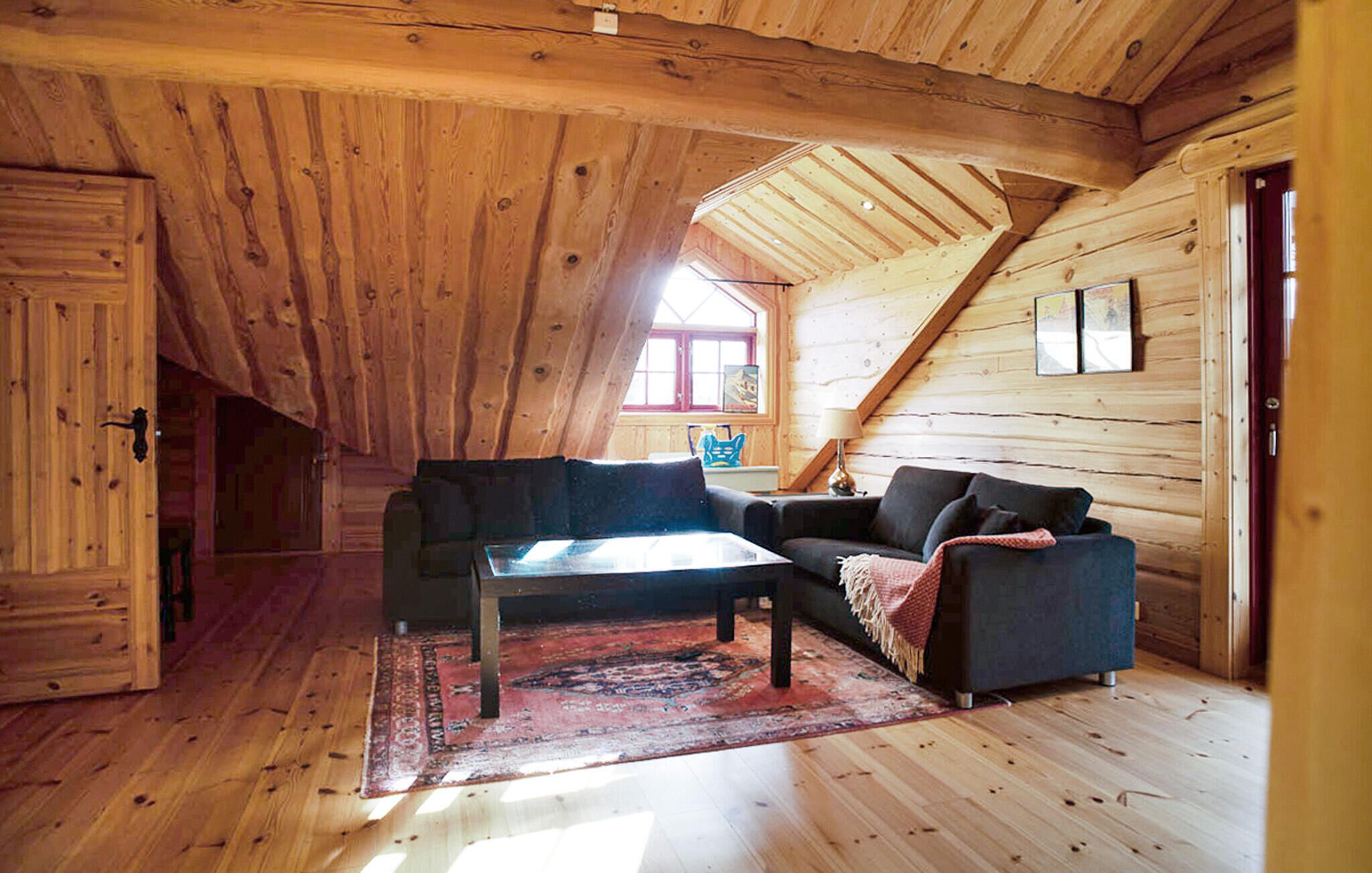 Un cuarto ático acogedor con paredes de madera y techo inclinado. Tiene un sofá de color oscuro, una pequeña mesa de centro y un tapete rojo con estampado.