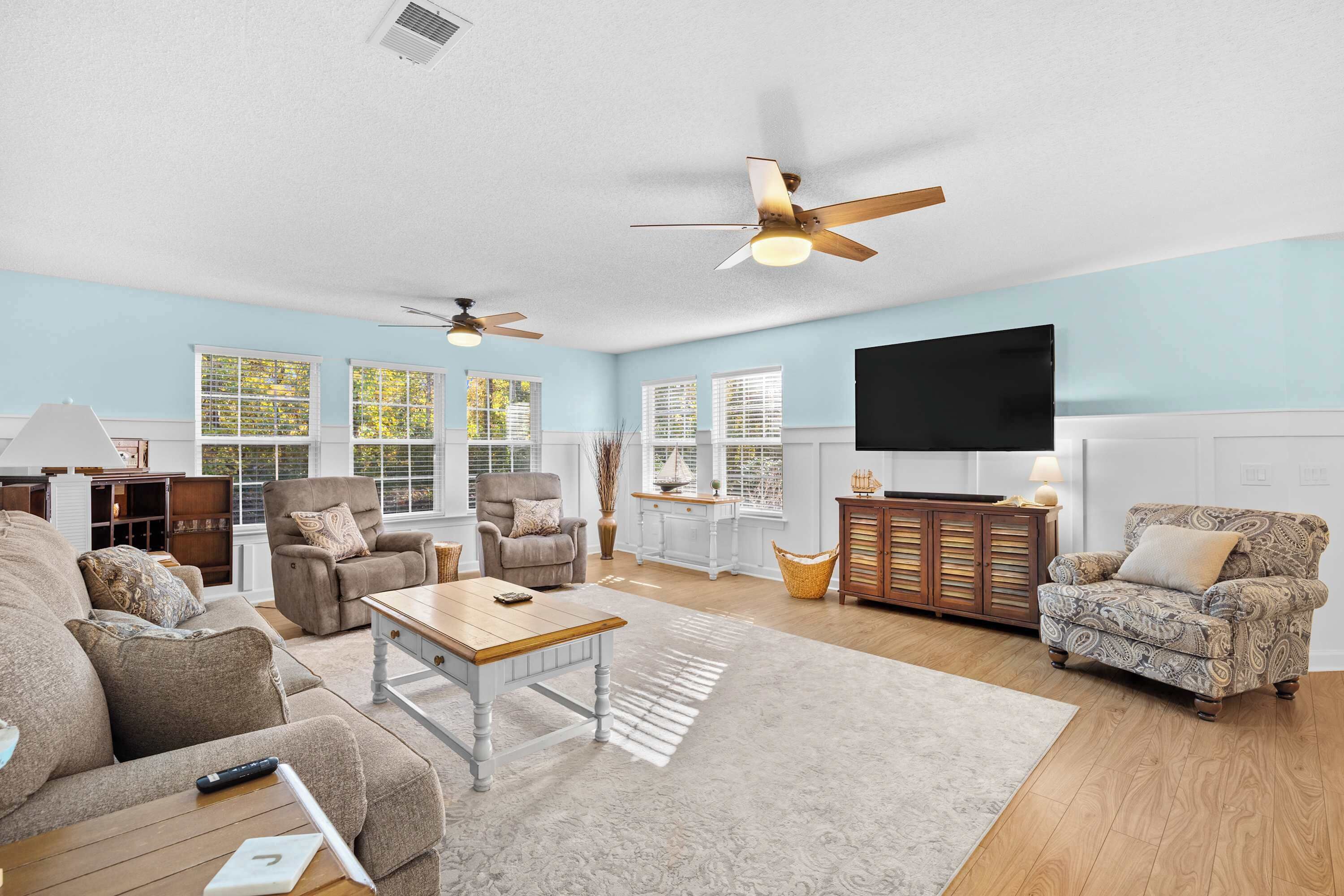 A spacious living room with a sofa, armchairs, a coffee table, and a fireplace. There are ceiling fans, a television, and a large window with blinds.