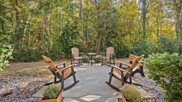 A circular outdoor seating area with four Adirondack chairs and a small round table, surrounded by a forested landscape. - Coastal Comfort with Golf, Pool, Pickle Ball, Tennis Courts (Murrells Inlet)