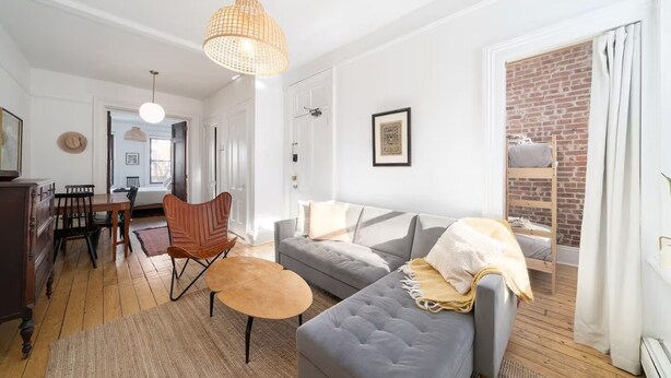 A modern living room with a grey sofa, wooden dining table, and chairs. There is a hanging light fixture and a brick wall in the background.