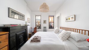 A bedroom with a large bed, a fireplace, a wooden dresser, and a view of the city through the windows.