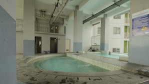 Une grande piscine intérieure, dont le sol et les murs sont recouverts de carrelage, est entourée d’un haut plafond laissant apparaître des tuyaux et d’un balcon doté de balustrades.