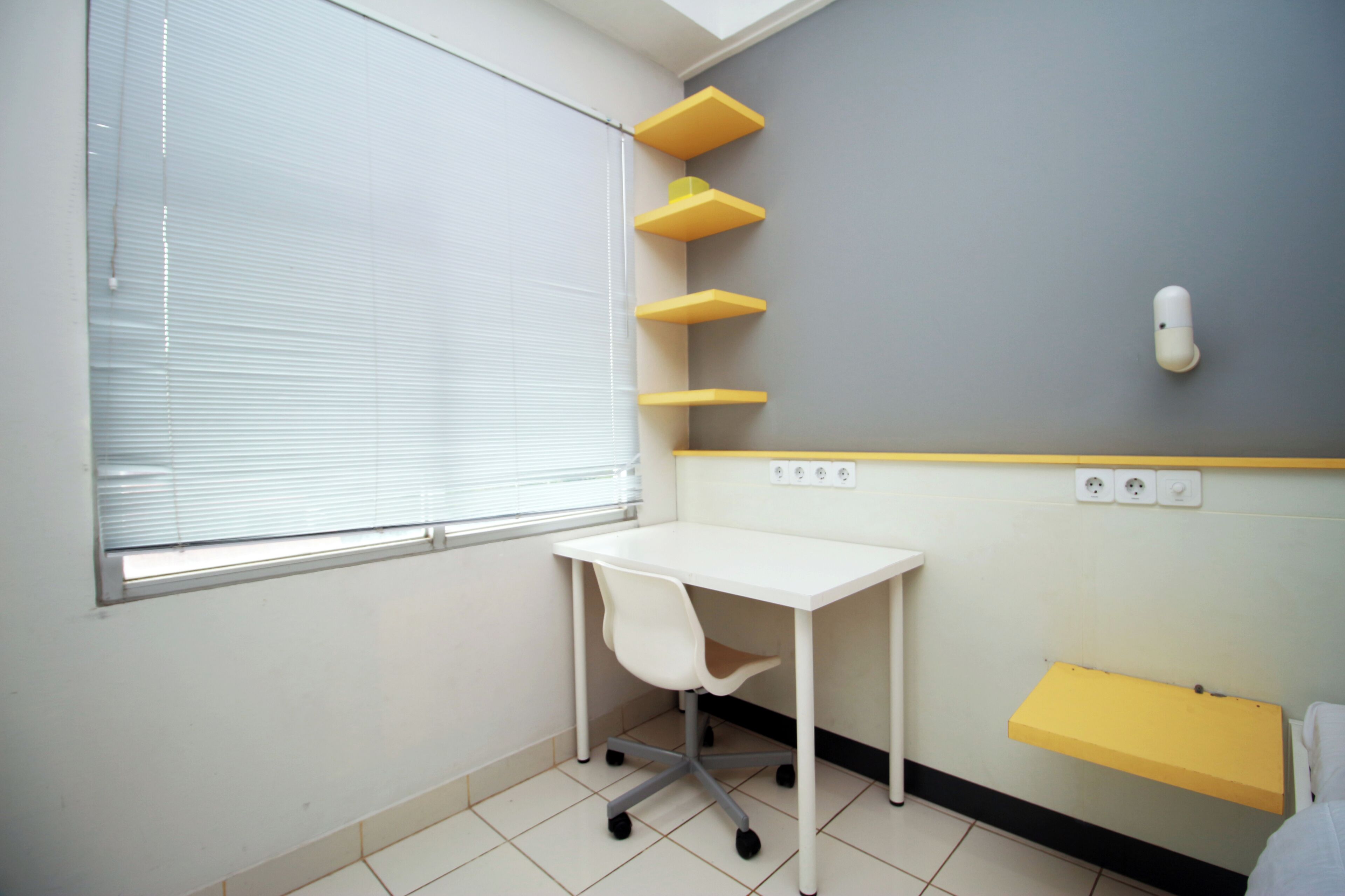 Een kleine, schone kamer met een wit bureau, een stoel, een raam met zonwering en een wandplank met een geel dienblad.