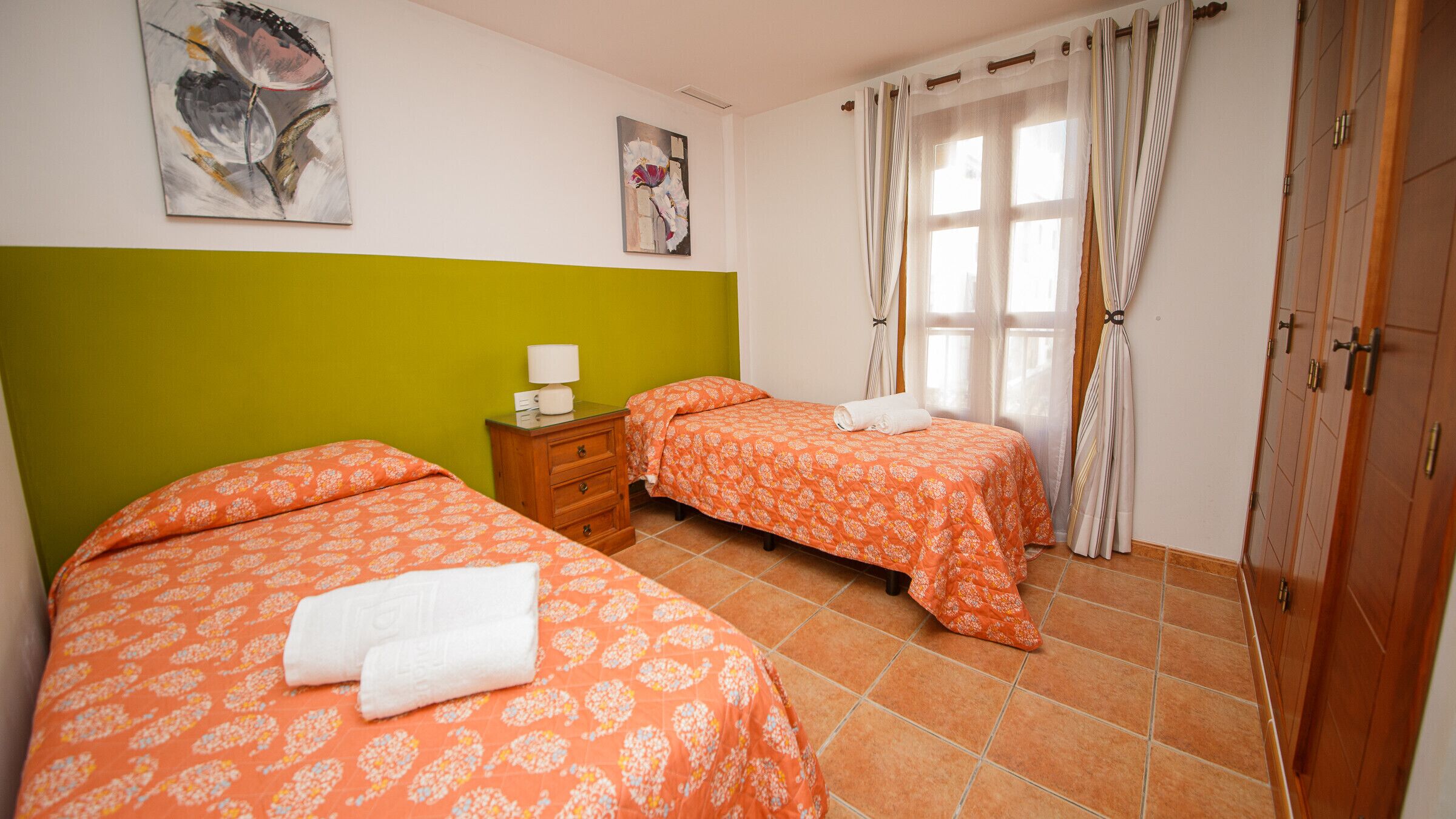 A bedroom with two beds, a nightstand, a lamp, and a window with curtains.