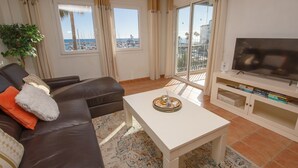 A living room with a brown leather sofa, a white coffee table, and a TV on a stand. There are two windows with curtains, a balcony with a view of the sea and buildings, and a plant on the left. - Marina Apartments 7.3.7 (Manilva)