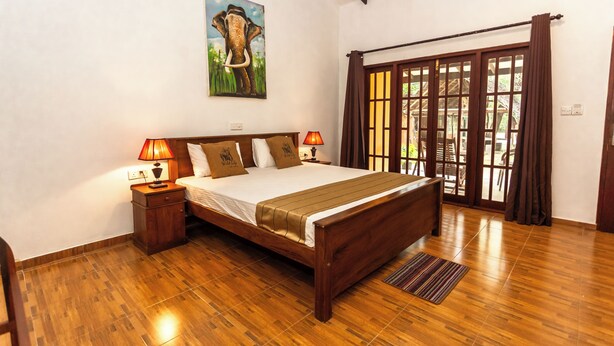 A wooden-framed bed with a white mattress and brown bedding, two bedside lamps, a wooden nightstand, a large window with brown curtains, and a painting of an elephant on the wall. - Elephant Path by Alpine Resorts (Habarana)