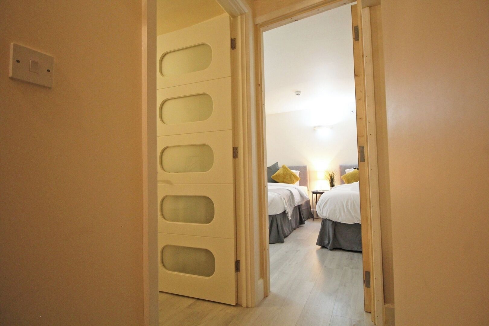 A hotel room with a wooden floor, a bed with a grey bedspread, and a bedside table with a lamp.