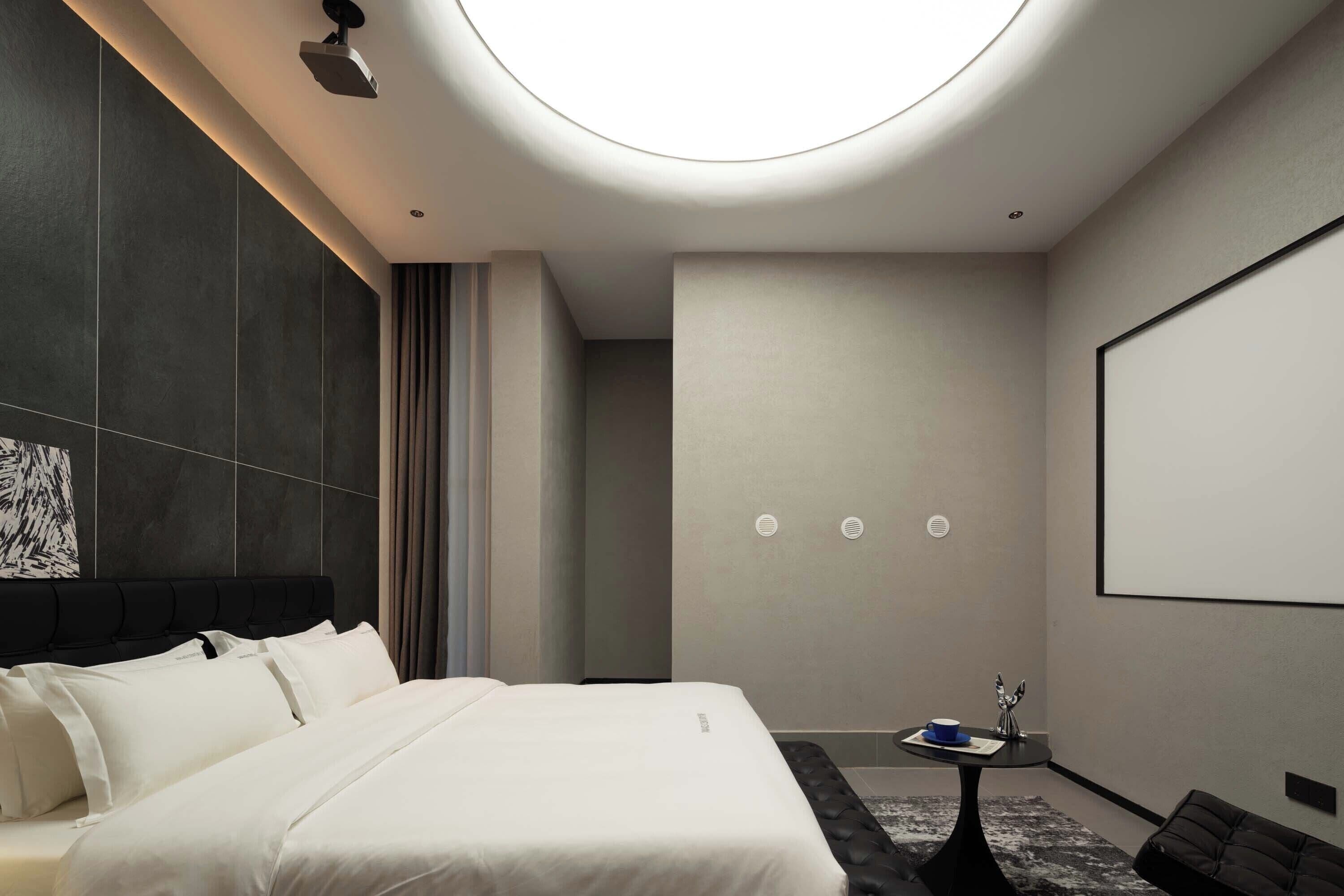 Ein modernes Hotelzimmer mit einem großen Bett, einem kleinen Tisch mit Tasse und Dekoration, einer Projektionsleinwand und einer Deckenlampe.