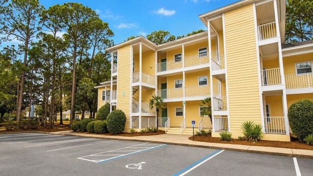 Un immeuble jaune à plusieurs étages, doté de balcons, un parking comprenant des places réservées aux personnes handicapées et des arbres environnants.