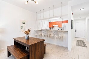 A modern kitchen with hanging macramé chairs, a wooden dining table with benches, and a white door. - Walk to Grand Blvd • Boho 2BR + Pool + Gym (Miramar Beach)