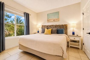 A bedroom with a large bed, two bedside tables, a window with curtains, a painting on the wall, and a door. - Walk to Grand Blvd • Boho 2BR + Pool + Gym (Miramar Beach)