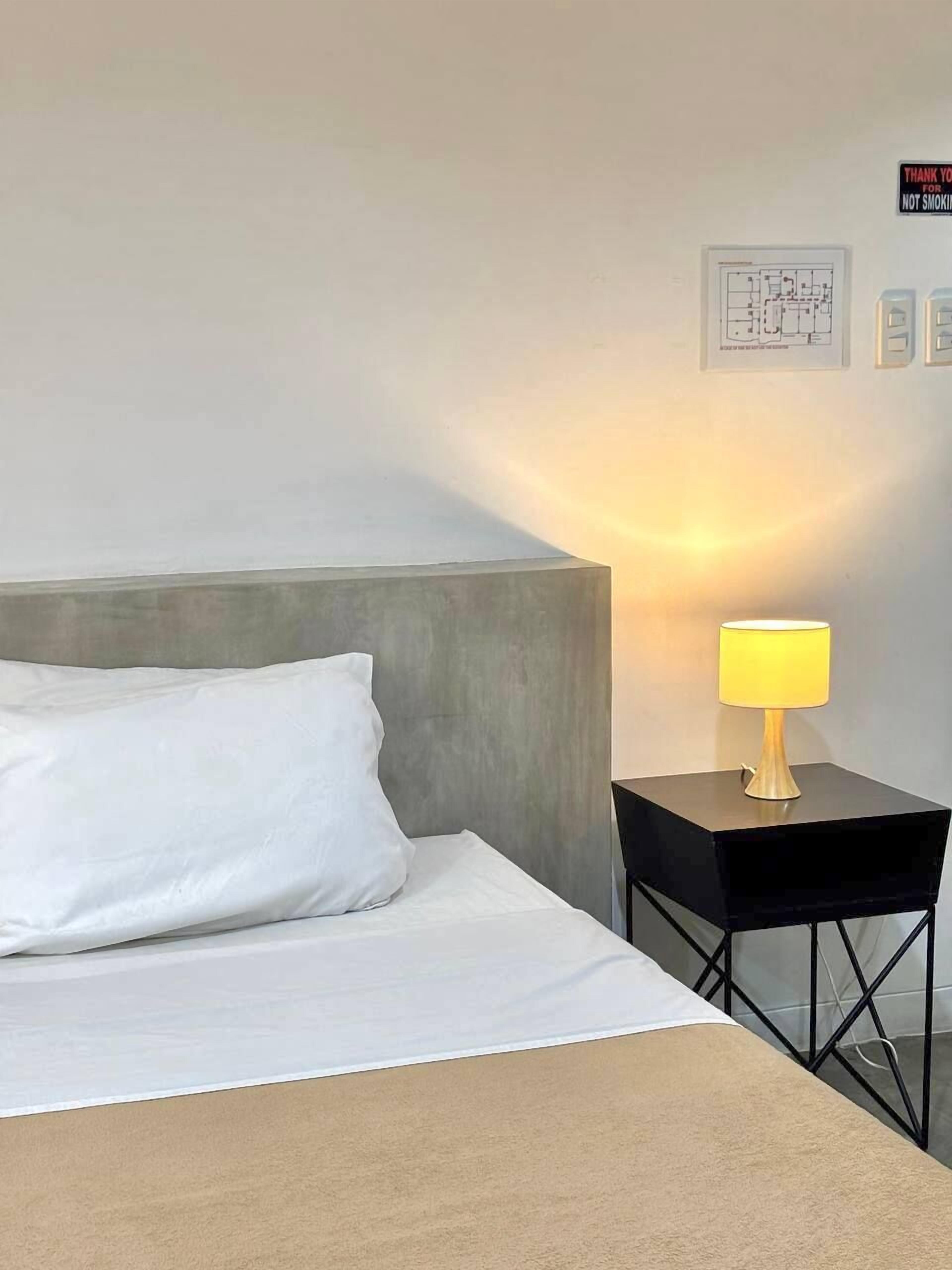 A neatly made bed with white bedding and a grey headboard, a small black nightstand with a yellow lamp, and a wall-mounted light fixture.