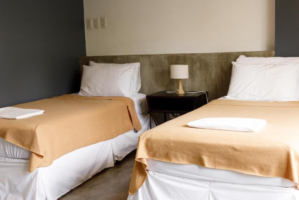 Une chambre d’hôtel avec deux lits, chacun recouvert d’une couverture beige et agrémenté d’oreillers blancs. Une petite table avec une lampe se trouve entre les deux lits.