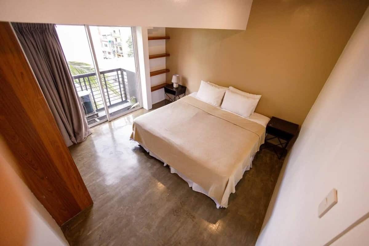 A bedroom with a bed, a nightstand, a lamp, and a view of a balcony.